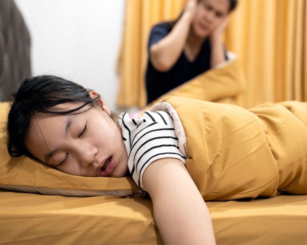 Niña roncando mientras duerme y mamá al fondo tapándose los oídos